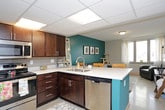 This single-floor unit offers a thoughtfully arranged layout that includes a living area, kitchen, multiple bedrooms, and two full bathrooms. The living room makes a strong visual impression with a bold teal accent wall, a gray sofa, and a coffee table, while large windows in the adjoining area flood the space with natural light and frame sweeping city skyline views. The kitchen is outfitted with dark espresso cabinetry, stainless steel appliances including a refrigerator, range, and microwave, light countertops with a tile backsplash, and direct access to the unit's entry door. The bedrooms are generously sized, each featuring large windows that offer expansive outdoor views of the surrounding cityscape and neighboring buildings. The rooms provide ample space for a bed, dresser, and dedicated study desk, making them well-suited for both rest and productivity. The unit includes two full bathrooms, both featuring dark espresso vanities with white countertops, mirrors with vanity lighting, and neutral gray walls. One bathroom includes a shower with a curtain, while the other features a more spacious layout with speckled tile flooring. Throughout the unit, drop-panel ceilings and light-toned walls contribute to a clean, functional aesthetic.