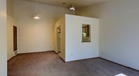 This single-floor residence offers a thoughtfully designed open layout that maximizes comfort and functionality. The main living area is anchored by soaring vaulted ceilings that lend an impressive sense of vertical space, complemented by a ceiling fan with a light fixture that ensures year-round comfort. Plush wall-to-wall carpeting in a neutral tone runs throughout, adding warmth underfoot and tying the interior together cohesively.

A defining architectural feature is the interior pass-through window cut into a partial dividing wall, which connects the living area to the kitchen. This design choice enhances the open feel of the home while maintaining a subtle separation between spaces. The kitchen, visible through the pass-through, features overhead cabinetry with under-cabinet lighting, offering a practical and well-lit workspace.

The crisp white walls and clean lines throughout the interior create a bright, airy ambiance that makes the space feel welcoming and easy to personalize. An arched passageway leads further into the home, hinting at additional rooms beyond. The overall aesthetic is simple and classic, providing a versatile canvas suitable for a variety of interior styles and furnishing choices.
