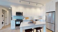 This single-floor space showcases a thoughtfully designed modern kitchen that combines clean aesthetics with practical functionality. The kitchen is defined by its crisp, handle-free white cabinetry that extends from floor to ceiling, maximizing storage while maintaining a seamless, minimalist appearance. A soft blue tile backsplash adds a subtle pop of color, creating a pleasing contrast against the all-white cabinetry and bright white quartz countertops.

The kitchen is equipped with stainless steel appliances, including a gas range, over-the-range microwave, and additional built-in appliances, delivering a cohesive and professional-grade cooking environment. A centrally positioned island with a white quartz surface provides generous prep space and features two dark wood bar stools, making it ideal for casual dining or entertaining.

Track lighting runs along the ceiling, directing focused illumination throughout the workspace and enhancing the kitchen's bright, airy atmosphere. Light wood-tone flooring complements the white palette, adding warmth to the otherwise contemporary design. Overall, the space projects a clean, open, and highly functional character well-suited to modern living.