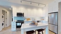 This single-floor space showcases a thoughtfully designed modern kitchen that combines clean aesthetics with practical functionality. The kitchen is defined by its crisp, handle-free white cabinetry that extends from floor to ceiling, maximizing storage while maintaining a seamless, minimalist appearance. A soft blue tile backsplash adds a subtle pop of color, creating a pleasing contrast against the all-white cabinetry and bright white quartz countertops.

The kitchen is equipped with stainless steel appliances, including a gas range, over-the-range microwave, and additional built-in appliances, delivering a cohesive and professional-grade cooking environment. A centrally positioned island with a white quartz surface provides generous prep space and features two dark wood bar stools, making it ideal for casual dining or entertaining.

Track lighting runs along the ceiling, directing focused illumination throughout the workspace and enhancing the kitchen's bright, airy atmosphere. Light wood-tone flooring complements the white palette, adding warmth to the otherwise contemporary design. Overall, the space projects a clean, open, and highly functional character well-suited to modern living.