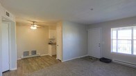 This single-floor unit presents a practical open-concept layout that connects the living area directly to the kitchen. The living space features wall-to-wall carpet, providing a comfortable underfoot feel, while the kitchen area transitions to wood-style vinyl flooring. Neutral gray walls throughout create a clean, cohesive aesthetic that complements the natural light filtering into the space.

The kitchen is equipped with white upper and lower cabinetry, offering ample storage, along with a countertop that appears to feature a stone-style finish. A doorway leads further into the unit from the kitchen area. The living area is equipped with a ceiling fan with integrated lighting, providing both air circulation and overhead illumination.

A baseboard heater is positioned along the living room wall, offering supplemental heating. The main entryway features a solid white door with a deadbolt, opening directly into the carpeted living space. A small entry mat is placed near the door. The space maintains a simple, functional design with clean lines and a neutral palette throughout.