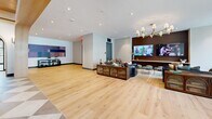 Located at 600 Kissimmee Drive in Plano, TX, this single-floor community space offers an expansive, thoughtfully designed interior that blends contemporary architecture with warm, inviting aesthetics. Wide-plank light oak hardwood flooring flows throughout, anchoring the open layout and connecting its many distinct zones seamlessly.

The main living area is bathed in natural light streaming through floor-to-ceiling steel-framed windows and glass doors that open to outdoor courtyard views. Multiple seating arrangements — including curved white sofas, retro-inspired lounge chairs, and intimate conversation groupings — create flexible social environments suited for both relaxed gatherings and vibrant community events.

A central kitchen or bar area features a bold checkerboard tile floor, dark stone countertops, and a striking coffered wood ceiling with pendant lighting, adding warmth and visual depth. Elegant arched doorways with wood detailing frame transitions between zones, providing architectural character throughout.

Gallery-style wall art is prominently displayed along open corridors, adding a refined cultural dimension to the space. A dedicated media lounge with a large wall-mounted TV, dark wood cabinetry, and a statement brass chandelier offers an intimate entertainment retreat. Recessed lighting throughout ensures a bright, welcoming ambiance at all hours, making this a truly dynamic and polished community hub.