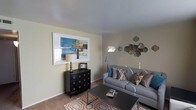 This single-story space presents a thoughtfully designed living area that blends contemporary style with everyday functionality. The living room serves as the focal point, featuring light-toned walls that create a bright and open atmosphere. A plush gray sofa anchors the seating area, dressed with an assortment of patterned throw pillows in neutral and teal tones that add visual interest. A sleek, dark rectangular coffee table sits atop a geometric-patterned area rug in warm orange and white tones, grounding the space with a pop of color. Against one wall, a bold black dresser with circular hardware provides both storage and a stylish surface for a classic table lamp, complemented by a large framed abstract artwork in blue and gold tones above it. A contemporary arc floor lamp adds ambient lighting to the room. Decorative metallic wall art in teal and bronze tones adorns the opposite wall, adding texture and dimension. The flooring appears to be a neutral carpet, contributing to the space's warm and comfortable feel. Overall, the living area exudes a balanced mix of modern design and inviting comfort.