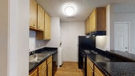 This single-floor, two-bedroom, one-bathroom residence in Little Rock, Arkansas offers a practical and well-organized layout. The galley-style kitchen is efficiently designed with warm oak cabinetry lining both walls, dark granite-look countertops, black appliances including a range and refrigerator, a stainless steel sink, and wood-look flooring. A stacked washer and dryer unit is conveniently housed within the kitchen area. The open living area features neutral-toned carpet, a ceiling fan with light fixture, and direct sightlines to the kitchen, creating a cohesive and connected common space. A sliding glass door in one area of the unit allows natural light to filter in and provides outdoor access. Both bedrooms are carpeted and finished with neutral walls and textured ceilings. Each room benefits from windows with blinds that bring in natural light, and at least one bedroom includes a walk-in closet for added storage. The bathroom is clean and straightforward, featuring a full tub-shower combination, toilet, and dark countertop vanity with wood-look flooring that ties back to the kitchen aesthetic. Overall, the residence offers a comfortable and functional living environment suitable for everyday living.