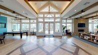 The Mason Freaux Park Clubhouse in Slidell, Louisiana is a thoughtfully designed single-story community gathering space that combines architectural elegance with functional amenities. The grand entrance immediately impresses with its dramatically vaulted ceiling, exposed wood beam trusses, and a striking geometric-patterned hardwood floor featuring contrasting inlays. Floor-to-ceiling windows and glass double doors flood the space with natural light while offering views of the surrounding community.

The open main hall transitions seamlessly into distinct activity zones. A lounge area features tufted seating, a teal shiplap accent wall with a mounted flat-screen TV, and decorative chandelier lighting. Adjacent to this is a dedicated game room with a full-size billiard table, dual wall-mounted TVs, exposed beam ceilings, and tall windows overlooking the exterior.

A kitchenette area provides white cabinetry, a sink, countertop workspace, and a decorative living wall of mounted planters, adding a fresh botanical element to the interior. The space also includes private office or meeting rooms enclosed by glass-panel partitions, maintaining an open, airy feel throughout. The overall design blends coastal-inspired colors, warm wood tones, and modern fixtures to create an inviting, upscale community environment.