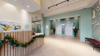 Located at 601 N Broad St in Philadelphia's vibrant North Broad corridor, this ground-floor lobby offers a welcoming and thoughtfully designed entry experience. The space is anchored by a curved, slatted-wood reception desk finished in warm natural tones, positioned prominently near the floor-to-ceiling glass entrance doors that flood the interior with abundant natural light. Soaring ceilings with exposed track lighting and a sculptural multi-arm pendant chandelier contribute to an open, airy ambiance throughout. The flooring consists of large-format neutral-toned tiles that extend seamlessly across the entire space. Sage green accent walls lend a calm, modern aesthetic, complemented by a striking large-scale abstract mural wallcovering that serves as a focal point along one wall. A pair of elevators, flanked by built-in wood bench seating and potted planters, provides access to upper floors. An extended hallway with a mailbox bank connects to additional building amenities. The lobby's design balances functional elements — including a secure entry vestibule and resident mailboxes — with polished, contemporary finishes, creating a refined and inviting first impression for residents and visitors alike.