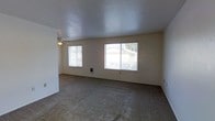 This single-floor space offers a straightforward, functional layout centered around a generously sized living area. The room features wall-to-wall carpeting underfoot and a textured popcorn ceiling overhead, lending a classic residential character to the interior. Two large windows along the exterior wall allow abundant natural light to fill the space, offering views of the surrounding neighborhood. Horizontal blinds on each window provide adjustable light control and privacy. A ceiling fan is installed overhead, offering both lighting and air circulation for year-round comfort. The neutral, light-colored walls create a clean and adaptable backdrop suited to a variety of furnishing styles. A wall-mounted heating unit is conveniently positioned beneath one of the windows, ensuring efficient climate control. The open, unobstructed floor plan of the living area provides flexibility in how the space can be arranged and utilized. Overall, the space presents a practical and livable environment with essential amenities in place, ready to be personalized to suit the needs of its occupants.