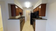 The kitchen is designed in a classic galley configuration, with parallel countertops and cabinetry lining both sides of a central walkway, creating an efficient and organized cooking environment. Rich, warm-toned wood cabinets extend from counter height to near the ceiling on both sides, providing generous storage space throughout. The countertops feature a dark granite-look surface that contrasts nicely with the lighter-colored walls, adding visual depth to the space.

On one side, a stainless steel sink is integrated into the counter, accompanied by a dishwasher beneath. The opposite side houses a black electric range and overhead microwave, keeping the cooking zone consolidated and practical. A full-size black refrigerator anchors the far end of the kitchen, completing the suite of appliances.

The light tile flooring brightens the space and complements the overall color palette. Overhead, a flush-mount ceiling light provides consistent illumination throughout the galley. A textured ceiling adds a subtle architectural detail. At the far end, a doorway leads to an adjacent utility or laundry area, enhancing the overall functionality of this well-organized kitchen layout.