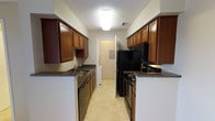 The kitchen is designed in a classic galley configuration, with parallel countertops and cabinetry lining both sides of a central walkway, creating an efficient and organized cooking environment. Rich, warm-toned wood cabinets extend from counter height to near the ceiling on both sides, providing generous storage space throughout. The countertops feature a dark granite-look surface that contrasts nicely with the lighter-colored walls, adding visual depth to the space.

On one side, a stainless steel sink is integrated into the counter, accompanied by a dishwasher beneath. The opposite side houses a black electric range and overhead microwave, keeping the cooking zone consolidated and practical. A full-size black refrigerator anchors the far end of the kitchen, completing the suite of appliances.

The light tile flooring brightens the space and complements the overall color palette. Overhead, a flush-mount ceiling light provides consistent illumination throughout the galley. A textured ceiling adds a subtle architectural detail. At the far end, a doorway leads to an adjacent utility or laundry area, enhancing the overall functionality of this well-organized kitchen layout.