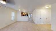This single-floor residence is designed with an open-concept layout that promotes a seamless flow between the main living areas. The living room is generously sized, featuring plush neutral-toned carpeting, a ceiling fan, and multiple windows fitted with blinds that allow natural light to fill the space. The exterior views visible through the windows offer glimpses of a neighboring brick building and surrounding environment.

A half-wall peninsula connects the living area to the kitchen, which is finished with warm wood-toned cabinetry on both upper and lower sections. The kitchen is equipped with black appliances, including a refrigerator, range, and dishwasher, complemented by light countertops and a double-basin sink. Wood-look flooring runs throughout the kitchen, providing a practical contrast to the carpeted living spaces. A window above the counter and additional windows along the walls bring in natural light.

The layout also includes multiple rooms accessible via a hallway, each featuring carpet flooring, ceiling fans, and windows. Several sets of double-door closets with paneled white doors are visible throughout, offering generous storage. The overall color palette is light and neutral, with white trim and walls contributing to a clean, cohesive aesthetic throughout the space.