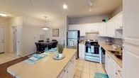 This single-story residence features a thoughtfully designed open-concept floor plan that seamlessly connects the main living areas. The living room is highlighted by vaulted ceilings, a ceiling fan with light fixture, and rich hardwood-style flooring. Teal accent curtains frame sliding glass doors that open to an outdoor space, filling the room with natural light.

The kitchen is well-equipped with white cabinetry, light countertops, stainless steel appliances including a refrigerator and range, and a tile floor. A peninsula with bar seating serves as both a functional workspace and casual dining area, while track lighting overhead provides ample illumination. An adjacent dedicated dining area accommodates a full dining set beneath a pendant light fixture.

The bedroom offers a calm retreat with plush carpeting, a ceiling fan, and multiple windows that allow natural light to flow in. The full bathroom features tile flooring, a vanity with generous counter space, and a soaking tub. Notably, a walk-in closet connects directly to the bathroom, providing substantial storage. The overall aesthetic is cohesive and bright, with neutral walls accented by consistent teal tones throughout.