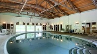 This indoor aquatic facility is defined by its impressive architectural design, centered around a soaring vaulted ceiling constructed from warm wood planking supported by exposed steel trusses. The structural framework overhead creates a dramatic, open feel while adding a rustic yet refined aesthetic to the space.

The generously sized pool occupies the heart of the facility, its calm, reflective water contributing to the tranquil atmosphere. The curved pool edges and smooth surrounding deck provide ample space for movement and poolside activities. Underwater lighting enhances the pool's visual appeal, casting a soft glow across the water's surface.

Along one wall, a row of windows allows natural light to filter in, offering views of the surrounding greenery outside. This connection to the outdoors further enhances the serene environment within. The white walls complement the warm tones of the ceiling, keeping the space feeling bright and airy.

Along the perimeter, dark-paneled changing room or locker room doors are evenly spaced, providing functional amenities for users. Potted plants are placed near the facility's edges, adding subtle natural accents. The overall ambiance is calm, clean, and well-suited for both recreational and therapeutic aquatic use.