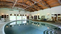 This indoor aquatic facility is defined by its impressive architectural design, centered around a soaring vaulted ceiling constructed from warm wood planking supported by exposed steel trusses. The structural framework overhead creates a dramatic, open feel while adding a rustic yet refined aesthetic to the space.

The generously sized pool occupies the heart of the facility, its calm, reflective water contributing to the tranquil atmosphere. The curved pool edges and smooth surrounding deck provide ample space for movement and poolside activities. Underwater lighting enhances the pool's visual appeal, casting a soft glow across the water's surface.

Along one wall, a row of windows allows natural light to filter in, offering views of the surrounding greenery outside. This connection to the outdoors further enhances the serene environment within. The white walls complement the warm tones of the ceiling, keeping the space feeling bright and airy.

Along the perimeter, dark-paneled changing room or locker room doors are evenly spaced, providing functional amenities for users. Potted plants are placed near the facility's edges, adding subtle natural accents. The overall ambiance is calm, clean, and well-suited for both recreational and therapeutic aquatic use.
