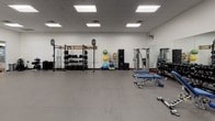 This single-floor fitness facility is designed to accommodate a broad range of workout disciplines within a spacious, open layout. The interior features white walls, a drop-panel ceiling with recessed fluorescent lighting, and durable gray rubber flooring throughout, creating a clean and functional environment.

The space is organized into distinct training zones. The cardio area includes a row of stationary cycling bikes, treadmills, and a stair climber, positioned near a glass partition wall that separates adjacent areas while maintaining visual openness. A wall-mounted flat-screen television is installed nearby for entertainment or instructional content.

The strength training zone is well-appointed with a power rack, barbell storage, weight plates, dumbbells, and a cable machine. Additional equipment includes adjustable benches, medicine balls, stability balls, and resistance training accessories stored on dedicated racks along the walls. Large mirrors line one wall, enhancing the sense of space and allowing users to monitor their form.

Wall-mounted fans provide air circulation, and the open floor plan allows ample room for functional training and free movement between equipment stations. The overall ambiance is practical and professional, with a well-organized arrangement that supports both individual training and group fitness use.