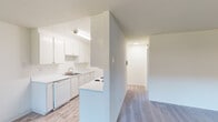 This single-floor unit presents a bright and clean interior with a consistently neutral color palette throughout. The galley-style kitchen is a functional highlight, featuring white upper and lower cabinetry offering generous storage, white countertops, a stainless steel sink, and a full suite of white appliances including an electric range, built-in microwave, refrigerator, and dishwasher. Light wood-look flooring runs through the kitchen and connects seamlessly to the entryway and hallway areas. The open living space transitions from wood-look flooring to plush carpeting, creating a comfortable, defined zone ideal for relaxing or entertaining. A large window fitted with vertical blinds allows natural light to fill the room while offering outdoor views of surrounding greenery. The bedrooms are generously sized, each finished with soft carpet and neutral walls, providing a clean canvas for personalization. A baseboard heater in one bedroom ensures year-round comfort. The bathroom is well-appointed with a wide vanity topped with a speckled countertop, a large mirror with overhead lighting, and a full tub and shower combination with clean white tile surround. The overall ambiance is light, airy, and move-in ready.