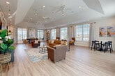 Located in Colorado Springs, CO, this single-floor community space offers a thoughtfully designed open-plan layout that seamlessly blends a living room and kitchen into one cohesive environment. Light wood-plank flooring flows throughout, unifying the space and enhancing its bright, welcoming atmosphere.

The expansive living area is anchored by multiple large windows dressed with patterned grommet curtains, flooding the room with natural light and offering views of the surrounding neighborhood. Recessed lighting and a tray ceiling detail add architectural interest overhead. Comfortable seating arrangements — including a caramel leather sofa, accent armchairs, and a casual high-top dining table — create distinct zones for relaxing, socializing, and gathering. A wall-mounted television above a chevron-paneled media console serves as a focal point on one end of the room.

The galley-style kitchen is a standout feature, finished with rich dark cabinetry, marble-look quartz countertops, and a bold navy accent wall that adds depth and contrast. Stainless steel appliances, a double sink, and ample counter space make it well-equipped for entertaining. A centered window brings in additional natural light, completing this functional yet stylish kitchen. Together, these spaces form a polished, inviting environment ideal for community use.