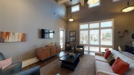 This single-floor communal space is defined by its striking architectural design, highlighted by dramatically vaulted ceilings that soar overhead and are accentuated by clerestory windows, allowing abundant natural light to fill the interior. The main lounge area is generously proportioned, featuring wood-look tile flooring, a mix of stylish seating arrangements, and a wall-mounted flat-screen television set atop a warm wood media credenza. Large floor-to-ceiling windows and glass doors frame views of an outdoor pool area, seamlessly connecting the interior to the exterior environment. Pendant lighting and drum-style fixtures complement the open, airy ambiance throughout. Structural columns delineate different zones within the open-plan layout, adding an element of architectural character. The kitchen is well-appointed, showcasing rich dark cabinetry that extends to the ceiling, light granite countertops, wood-look tile flooring, stainless steel appliances including a refrigerator and built-in microwave, and ample workspace. A secondary kitchen area features a double sink, additional cabinetry, and a window with natural light. The overall palette of warm gray, taupe, and earthy tones creates a welcoming and cohesive atmosphere throughout the space.