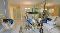 This single-floor space exudes a refined, eclectic elegance through its thoughtfully curated interior design. The open-plan layout connects the living, dining, and kitchen areas in a cohesive flow, creating a sense of spaciousness and ease of movement throughout.

The living area is anchored by a glass-top table adorned with a striking sculptural peacock figurine, lending an artistic, decorative focal point to the room. White upholstered seating and ornate metalwork accents complement the space's sophisticated aesthetic. Floor-to-ceiling mirrored wall panels line one side of the room, visually expanding the interior and adding depth and luminosity.

The dining area features classically styled chairs with intricately carved frames and patterned upholstery, arranged around a formal dining table beneath a warm chandelier. A built-in display shelf nearby showcases decorative dishware and glassware, adding a curated, personal touch.

The kitchen, visible through a pass-through opening, is accented by tall teal glass bottles that introduce a bold pop of color against neutral cabinetry. Light-toned tile flooring runs throughout, tying the spaces together with a clean, consistent finish. Warm ambient lighting from flush-mount fixtures and the chandelier creates a welcoming and polished atmosphere throughout this distinctive home.
