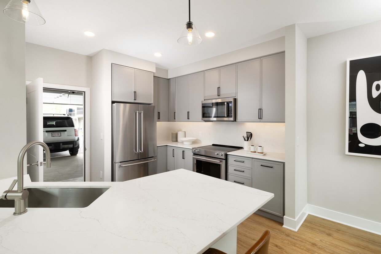 Signature Collection kitchen with stainless steel appliances, white quartz countertop, grey cabinetry, white quartz backsplash, hard surface flooring, and attached garage - Avalon Wayne