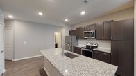 This single-floor residence offers a thoughtfully designed open-concept layout that seamlessly connects the kitchen, dining, and living areas. The kitchen serves as the centerpiece of the home, showcasing a generous granite-topped island with an integrated sink, rich dark-brown cabinetry, a classic white subway tile backsplash, and a full suite of stainless steel appliances including a refrigerator, electric range, and over-the-range microwave. Wood-look flooring runs throughout the kitchen and main living corridors, lending warmth and continuity to the space.

The living area is bright and open, with plush neutral carpeting and multiple windows fitted with blinds that invite natural light while maintaining privacy. A door provides direct access to the outdoors, further enhancing the airy feel of the space. The bedroom continues the carpeted comfort, featuring a ceiling light fixture and a well-sized window with views of the surrounding exterior.

The bathroom is well-appointed with a tub-shower combination, brushed nickel fixtures, a curved shower rod, and dark vanity cabinetry topped with granite counters that echo the finishes found throughout the home. Neutral wall tones and clean white trim unify the design, creating a cohesive and modern aesthetic from room to room.