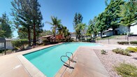 This single-story property offers a welcoming outdoor environment centered around a generously sized swimming pool. The pool area is a standout feature, showcasing clean, turquoise water with a classic pool entry ladder and a spacious concrete deck ideal for lounging or entertaining. The surrounding landscape is lush and well-maintained, with tall palm trees and mature evergreens providing both shade and a sense of seclusion. A wrought iron fence encloses the pool area, adding a layer of safety while maintaining an open, airy feel. Beyond the fencing, additional residential structures are visible among the greenery, suggesting a community-oriented setting. The abundant natural light and clear blue sky overhead enhance the bright, inviting atmosphere of the outdoor space. The combination of tropical palms, dense tree coverage, and manicured grounds creates a resort-like ambiance that is both relaxing and visually appealing. Overall, this property's outdoor amenities make it a compelling space for those who value leisure, natural beauty, and a vibrant outdoor lifestyle.