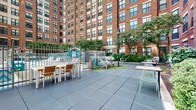 Located at 8204 Baltimore Ave in College Park, Maryland, this outdoor amenity courtyard is nestled within the interior of a multi-story brick residential building. The space is defined by its striking architectural backdrop — a tall, classic red-brick façade with large green-trimmed windows that wraps around the courtyard on multiple sides, creating an enclosed and intimate outdoor environment.

At the heart of the space is a generously sized outdoor swimming pool with clear, blue water, enclosed by a white metal fence with a gated entry. The pool area is complemented by a pergola structure and lined with lounge chairs and sun loungers, providing ample space for relaxation and recreation under open skies.

Surrounding the pool, the courtyard is finished with a combination of large-format slate-toned tile pavers and wooden deck sections, offering distinct zones for outdoor seating and socializing. Outdoor dining tables with chairs and Adirondack-style seating in teal and white are thoughtfully arranged throughout, alongside classic lamp posts that add a charming, pedestrian-friendly ambiance.

Mature trees and lush green landscaping are interspersed throughout the courtyard, softening the urban architecture and providing natural shade. The overall design balances an active outdoor lifestyle with relaxed gathering spaces, making this courtyard a well-appointed amenity for the surrounding community.