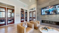 This single-floor leasing office and lounge is a thoughtfully designed space that balances professional functionality with an inviting, upscale ambiance. The layout is divided into two distinct yet connected areas: a welcoming lounge and a dedicated office workstation zone.

The lounge area immediately captures attention with its dramatic floor-to-ceiling stone tile accent wall, which serves as the backdrop for a sleek linear gas fireplace and a wall-mounted flat-screen television. Tall ceilings with crown molding lend an open, airy feel, while rich mahogany-framed glass entry doors with transom windows create a grand first impression. Plush, channel-tufted caramel leather armchairs arranged around a round white marble coffee table offer a comfortable and stylish setting for guests.

The office area features a large, contemporary wood-toned L-shaped desk equipped with dual workstations, providing ample space for staff to assist visitors. A vibrant blue subway tile backsplash with cabinetry and a sink adds a pop of color to this functional zone. Artwork adorns the walls, enhancing the professional yet welcoming atmosphere.

Throughout, light wood-look tile flooring unifies the space and complements the neutral color palette. A generous bank of large windows floods the interior with natural light and offers views of the surrounding residential community, making this space both practical and visually engaging.