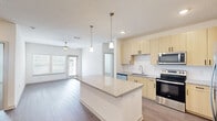 This single-story residence offers a thoughtfully organized layout across two bedrooms, two bathrooms, a living room, kitchen, dedicated laundry room, and two hallways connecting the spaces. The open-concept kitchen and living area forms the heart of the unit, featuring light wood-toned cabinetry, granite-look countertops, a central island with pendant lighting, and stainless steel appliances including a range, microwave, and refrigerator. Wood-look flooring flows throughout the main living areas, while a ceiling fan and large windows allow natural light to fill the living room.

Both bedrooms are carpeted and finished with neutral tones, each featuring windows with blinds and ceiling fans for added comfort. The primary bedroom includes a spacious walk-in closet equipped with wire shelving. The bathrooms mirror the kitchen's design aesthetic, with light wood cabinetry, granite-look countertops, and dark-framed mirrors. The dedicated laundry room houses a full-size washer and dryer alongside overhead wire shelving for additional storage. Throughout, the unit maintains a clean, contemporary aesthetic with a consistent neutral color palette and modern fixtures.