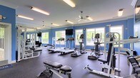 This fitness center offers a thoughtfully designed workout space finished with bold blue accent walls and dark rubber flooring that provides both durability and comfort underfoot. Large windows line the walls, allowing abundant natural light to fill the room while offering views of the surrounding outdoor greenery, creating an energizing atmosphere for exercise. Ceiling fans are mounted overhead alongside bright fluorescent lighting, ensuring the space remains well-ventilated and evenly illuminated throughout the day. A mounted flat-screen television is positioned on the wall, providing entertainment during workouts. The cardio area is well-stocked with multiple treadmills, elliptical machines, and upright exercise bikes, arranged to maximize floor space and ease of use. Strength training is supported by a selection of free weights, a weight bench, and cable resistance equipment. Full-length mirrors along one wall visually expand the space while allowing users to monitor their form during exercise. The overall layout is open and functional, providing ample room to move between stations comfortably, making this fitness center a practical and inviting facility for a well-rounded workout routine.