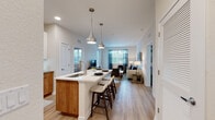 This well-appointed single-floor residence showcases a thoughtfully designed open-concept layout that seamlessly connects the kitchen, dining, and living areas. The kitchen is a standout feature, offering crisp white upper cabinetry paired with warm wood-toned lower cabinets, a large center island with a white quartz countertop, an undermount sink, and seating for casual dining. Sleek stainless steel appliances, under-cabinet lighting, and a classic subway tile backsplash complete the space. Elegant teardrop pendant lights hang above the island, adding a modern touch. Light wood-style flooring flows consistently throughout the residence, enhancing the sense of openness and continuity. The living area is bright and welcoming, benefiting from large windows dressed with teal accent curtains that invite natural light and provide outdoor views. The bedrooms are comfortably sized, each featuring windows with ample natural light and access to well-organized closet space, including a spacious walk-in closet with wire shelving. The bathroom is finished with a warm wood vanity, a white countertop, an illuminated frameless mirror, and matching light flooring. Clean lines, neutral tones, and contemporary finishes define the overall aesthetic of this modern residence.