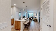 This well-appointed single-floor residence showcases a thoughtfully designed open-concept layout that seamlessly connects the kitchen, dining, and living areas. The kitchen is a standout feature, offering crisp white upper cabinetry paired with warm wood-toned lower cabinets, a large center island with a white quartz countertop, an undermount sink, and seating for casual dining. Sleek stainless steel appliances, under-cabinet lighting, and a classic subway tile backsplash complete the space. Elegant teardrop pendant lights hang above the island, adding a modern touch. Light wood-style flooring flows consistently throughout the residence, enhancing the sense of openness and continuity. The living area is bright and welcoming, benefiting from large windows dressed with teal accent curtains that invite natural light and provide outdoor views. The bedrooms are comfortably sized, each featuring windows with ample natural light and access to well-organized closet space, including a spacious walk-in closet with wire shelving. The bathroom is finished with a warm wood vanity, a white countertop, an illuminated frameless mirror, and matching light flooring. Clean lines, neutral tones, and contemporary finishes define the overall aesthetic of this modern residence.