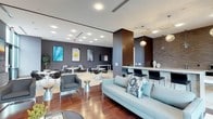 This single-floor contemporary lounge space is thoughtfully designed to blend comfort and modern aesthetics in an open, welcoming layout. The space is anchored by rich hardwood flooring in the main seating area, which transitions seamlessly into lighter tile flooring near the dining and bar zones, creating a natural delineation between functional areas.

The lounge area features a plush light-blue sectional sofa accented with decorative throw pillows, complemented by sleek modern armchairs and metallic side tables. A stylish bar counter with black high-back stools serves as a central gathering point, illuminated by elegant pendant lights that hang from a clean white ceiling lined with recessed lighting throughout.

The dining area offers multiple round tables paired with contemporary molded chairs in a mix of black and white, arranged beneath a dramatic charcoal accent wall adorned with a curated gallery of large-scale botanical and abstract artwork. A warm wood-paneled column provides architectural depth and visual contrast against the cool-toned walls.

The overall ambiance is polished and inviting, combining warm materials like wood and soft upholstery with modern finishes and purposeful lighting to create a versatile space suited for both relaxed socializing and casual gatherings.
