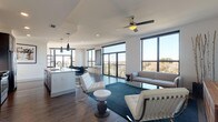 This stylish 2-bedroom, 2.5-bathroom residence offers a thoughtfully designed open-concept layout with contemporary finishes throughout. The expansive living area is anchored by large floor-to-ceiling windows framed in sleek black, flooding the space with natural light and offering sweeping outdoor views. A ceiling fan overhead and rich hardwood-style flooring add warmth and comfort to the living room, which flows seamlessly onto a private balcony.

The kitchen is a standout feature, showcasing grey cabinetry, a white quartz waterfall island with seating for three, stainless steel appliances, and elegant pendant lighting. A tasteful tile backsplash and ample storage complete this well-appointed space.

Both bedrooms are generously sized, each with direct balcony access through black-framed glass doors, offering bright views of the surrounding landscape and cityscape. The primary bathroom features marble-look tile flooring, a dark-cabinetry vanity with a white countertop, and a walk-in closet. The second full bathroom mirrors this refined aesthetic with clean white surfaces and a tiled shower surround. A convenient half bath rounds out the layout. Throughout, the design maintains a cohesive modern palette of whites, greys, and natural wood tones.