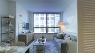 This single-floor space presents a well-designed living area centered around a striking set of large, floor-to-ceiling windows that frame views of the surrounding urban cityscape, flooding the interior with abundant natural light. The living room is thoughtfully arranged, featuring a plush gray sofa accented with decorative pillows in muted tones of blue and yellow, positioned to take full advantage of the expansive windows. A sleek glass-top coffee table rests atop a patterned area rug, adding texture and visual interest to the space. Opposite the seating area, a warm wood media console houses a wall-mounted flat-screen television, complemented by carefully arranged decorative accents including a small floral arrangement. A floor lamp with a soft white shade provides warm ambient lighting alongside the generous natural light. The walls are kept clean and neutral, with a single large-scale art piece adding a subtle artistic touch. The overall ambiance is calm and contemporary, blending functional urban living with an inviting, curated aesthetic that maximizes both comfort and style within its compact footprint.