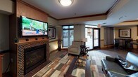 This single-floor community amenity space is thoughtfully designed to offer both comfort and functionality across several distinct areas. The lounge area serves as the heart of the space, anchored by a gas fireplace framed in decorative tile beneath a wall-mounted flat-screen television. Warm wood crown molding and a multi-toned area rug add richness to the interior, while large floor-to-ceiling windows and a glass door flood the room with natural light and provide views of the outdoor surroundings. Comfortable seating, including armchairs and a sofa arranged around a glass-topped coffee table, creates an inviting environment for socializing or relaxing.

A dedicated quiet seating nook offers a more intimate retreat, furnished with upholstered chairs set against textured neutral walls accented with a classic chair rail. Decorative art and a frosted-panel window complete the refined atmosphere of this secondary lounge area.

The kitchen is generously proportioned and outfitted with rich wood cabinetry, granite countertops, a stainless steel microwave, and a full sink. A raised bar counter with seating provides additional gathering space, while pendant lighting overhead adds warm ambiance. Tile flooring runs throughout the kitchen, and an adjacent dining area with table and chairs rounds out the space, making it well suited for community events and catered gatherings.