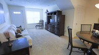 This single-floor space presents a comfortable and traditionally styled living area with a practical, well-organized layout. The room is finished with wall-to-wall plush carpeting in a warm neutral tone, contributing to a cozy and inviting atmosphere throughout.

The living area is generously proportioned, accommodating a large, dark wood entertainment center that serves as the room's focal point. The unit features built-in shelving, cabinet storage, and a central television bay, offering both functional media storage and display space. A classically styled accent chair with an upholstered floral pattern and matching ottoman adds a refined, traditional character to the space.

Natural light filters into the room through a window along the far wall, brightening the interior and lending an airy quality to the overall ambiance. The angled ceiling detail toward one end of the room adds architectural interest and dimension.

A partial view of a dining area is visible to the right, suggesting an open or semi-open floor plan that connects the living and dining spaces. The textured ceiling and neutral wall tones provide a clean, understated backdrop that complements the dark wood furnishings throughout the space.