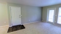This single-story space presents a straightforward and functional layout with a welcoming entryway that transitions seamlessly into the main living area. The entry features dark hardwood-style flooring, providing a durable and easy-to-clean surface near the front door, which is equipped with multiple security locks for added peace of mind. The living area is finished with light-colored carpet, contributing to a warm and comfortable atmosphere underfoot. Walls are painted in a soft, muted sage green tone that lends the space a calm and neutral ambiance, making it highly adaptable to a variety of furnishing styles and décor preferences. White baseboards provide clean contrast against the flooring and walls, adding a crisp, finished look throughout. The standard ceiling height and open wall space offer ample flexibility for furniture placement and personalization. Electrical outlets are conveniently positioned along the walls, and an additional doorway visible in the background suggests access to further rooms within the unit. Overall, the space is clean, well-maintained, and move-in ready.