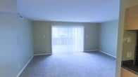 This single-floor space offers a clean and functional layout designed for comfortable everyday living. The living area is a standout feature, bathed in natural light streaming through a large sliding glass door dressed with vertical blinds, creating an airy and welcoming atmosphere. The neutral-toned walls complement the plush wall-to-wall carpeting underfoot, lending a soft, cohesive aesthetic to the space. The room is generously proportioned, providing ample space for a variety of furniture arrangements to suit different lifestyle needs. A floor vent along the baseboard ensures efficient climate control throughout the area, while the smooth white ceiling enhances the sense of openness. The sliding glass door not only floods the room with daylight but also provides direct access to an outdoor area, adding to the overall appeal of the space. The overall ambiance is understated and versatile, offering a blank canvas well-suited for personalization. With its emphasis on natural light, open layout, and practical finishes, this space provides a comfortable and inviting environment.