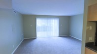 This single-floor space offers a clean and functional layout designed for comfortable everyday living. The living area is a standout feature, bathed in natural light streaming through a large sliding glass door dressed with vertical blinds, creating an airy and welcoming atmosphere. The neutral-toned walls complement the plush wall-to-wall carpeting underfoot, lending a soft, cohesive aesthetic to the space. The room is generously proportioned, providing ample space for a variety of furniture arrangements to suit different lifestyle needs. A floor vent along the baseboard ensures efficient climate control throughout the area, while the smooth white ceiling enhances the sense of openness. The sliding glass door not only floods the room with daylight but also provides direct access to an outdoor area, adding to the overall appeal of the space. The overall ambiance is understated and versatile, offering a blank canvas well-suited for personalization. With its emphasis on natural light, open layout, and practical finishes, this space provides a comfortable and inviting environment.