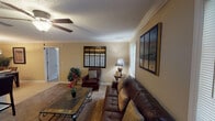 This single-floor residence offers a thoughtfully designed layout with comfortable, functional living spaces throughout. The living room serves as the heart of the home, featuring warm neutral walls, plush carpeting, crown molding, and a ceiling fan with light fixture. Multiple windows allow natural light to fill the room, complementing the cozy seating arrangement and decorative wall art. The space flows seamlessly into a dedicated dining area, which is highlighted by a pendant light fixture overhead and direct access to the kitchen. The kitchen is outfitted with wood cabinetry, dark granite-style countertops, tile flooring, and black appliances including a range and microwave. The primary bedroom is a serene retreat with carpeted floors, a window overlooking greenery, and direct access to an en-suite bathroom. The bathroom features warm wood cabinetry, a granite-topped vanity, a large mirror, tile flooring, and a separate toilet and tub area. A second bedroom mirrors the same comfortable aesthetic with plush carpeting and a textured ceiling. A walk-in closet is also accessible from the bedroom area. Throughout the residence, consistent warm beige tones, crown molding details, and textured ceilings create a cohesive and inviting ambiance.