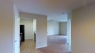 This single-floor residence showcases a clean, open layout designed for comfortable everyday living. The main living area is characterized by warm hardwood-style flooring that runs throughout, neutral-toned walls, and vaulted ceilings that lend a sense of openness and airiness to the space. Natural light filters in through a glass-panel door, brightening the living area and enhancing the overall welcoming ambiance.

The open-concept design allows for a seamless flow between the living area and the kitchen, which is visible through a wide passageway. The kitchen is equipped with dark appliances, including a full-size refrigerator, and features light-toned cabinetry and countertops, creating a functional and practical cooking space. White baseboards provide clean contrast against the neutral walls throughout the residence.

The layout is thoughtfully arranged, with clear sightlines from the kitchen through to the main living area, making the space feel connected and cohesive. The vaulted ceiling in the living area adds architectural interest, setting it apart from more conventional flat-ceiling designs. Overall, the residence presents a tidy, move-in-ready environment with a neutral palette that offers flexibility for a variety of interior styles and furnishings.
