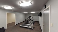 Located at 2233 W Jefferson St in Kokomo, Indiana, this fitness center is a single-floor facility designed to support a well-rounded workout experience. The space is organized into distinct zones, accommodating both cardiovascular training and strength work within a practical, functional layout.

The cardio area features multiple treadmills, elliptical trainers, and a stationary recumbent bike, each positioned on protective rubber floor mats atop durable carpet. Two wall-mounted flat-screen televisions are installed along one wall, allowing users to stay entertained during cardio sessions. A wall clock keeps users mindful of their workout time.

The strength training zone houses a multi-station cable and weight machine alongside a dedicated workout bench, free weights, and a stability ball, offering a variety of resistance training options. Protective rubber mats are placed beneath the equipment to safeguard the flooring. Motivational fitness posters are displayed along the walls, adding a purposeful atmosphere to the space.

Throughout the facility, flush-mount circular light fixtures provide consistent, bright overhead illumination. The textured ceilings, neutral gray walls, and dark carpeting create a clean, no-frills environment focused on functionality. A separate adjoining room offers additional space within the facility.