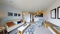 This single-floor space showcases a thoughtfully designed open-concept layout that integrates the living and kitchen areas into one cohesive, comfortable environment. The living area is generously sized, featuring soft carpeting accented by a stylish geometric-patterned area rug. A neutral color palette of cool grays and warm whites flows throughout, creating a calm and welcoming ambiance. The space is furnished with a light-toned sofa adorned with colorful accent pillows and complemented by a patterned accent chair, offering ample seating centered around a sleek rectangular coffee table. Wall art adds subtle decorative character to the space. The kitchen, visible from the living area, features dark cabinetry, a raised breakfast bar, pendant lighting, and modern appliances, making it both stylish and functional. Recessed lighting in the kitchen and ceiling-mounted fixtures provide consistent illumination throughout. A hallway leading off the main living area provides access to additional rooms, including what appears to be a laundry area with stacked units visible. The durable wood-look flooring in the kitchen and entryway transitions smoothly into the carpeted living space, tying the design together cohesively.