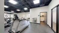 Located at 1000 Washington Ave in St. Louis, Missouri, this fitness center offers a well-equipped, single-floor layout designed to support a comprehensive workout experience. The main exercise area is generously sized, featuring durable rubber flooring throughout that provides both safety and comfort underfoot. The space is outfitted with a robust selection of True-brand cardio machines, including treadmills and recumbent bikes with interactive digital displays, alongside a cable machine and a variety of strength-training stations with adjustable benches. A large wall-mounted mirror along one side of the main floor visually expands the space and allows users to monitor their form during workouts. Structural columns are integrated tastefully into the layout, lending an open, loft-like character to the interior. The black drop-panel ceiling with recessed panel lighting creates a sleek, modern aesthetic while providing even, bright illumination throughout. A stainless steel dual drinking fountain is conveniently positioned near the bathroom entrance. The bathroom provides essential facilities to complete the experience. The overall ambiance is clean, contemporary, and functional, making this fitness center a practical and well-appointed amenity.