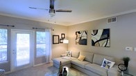This single-floor space presents a well-appointed living area designed with a clean, contemporary aesthetic. The room is finished with neutral beige walls that create a warm, inviting atmosphere, complemented by wall-to-wall carpeting that adds comfort underfoot. Crown molding runs along the ceiling, lending a refined architectural detail to the space.

A ceiling fan with a light fixture is centrally positioned, providing both ventilation and ambient lighting. Natural light enters through a large window dressed with sheer white curtains and a curtain rod, offering a bright, airy feel while maintaining privacy. An adjacent door with glass panels further enhances the flow of natural light into the room.

The living area accommodates a spacious L-shaped sectional sofa in a light gray upholstery, accented with decorative pillows in teal and yellow tones. A glass-top coffee table sits in front, adding a sleek, modern touch. The walls are adorned with framed abstract artwork in a blue, black, and cream palette, contributing visual interest and a cohesive modern style throughout the space. A floor lamp provides additional task lighting, completing the functional and stylish layout.