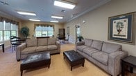 This single-floor communal space is thoughtfully designed to foster comfort and social interaction. The main lounge area is generously proportioned, featuring plush gray sofas and tufted leather ottomans arranged to encourage conversation and relaxation. Recessed panel lighting in the ceiling provides consistent, even illumination throughout, while crown molding along the upper walls adds a refined architectural touch. Warm beige carpeting and neutral-toned walls create an inviting, understated ambiance.

A dedicated dining nook adjacent to the lounge offers an intimate setting with a classic round wood table and wrought iron chairs. Large windows dressed with ornate valances and blinds flood this area with natural light and offer views of the surrounding greenery, enhancing the sense of openness and connection to the outdoors.

The kitchenette is fully equipped for practical use, featuring warm wood cabinetry, a double sink, a full-size refrigerator, dishwasher, microwave, and countertop coffee maker. Light hardwood-style flooring in this area provides a clean, easy-to-maintain surface. A flat-screen television is also available in the lounge, adding a recreational element to the space. Framed artwork on the walls and tasteful decorative accents throughout contribute to the space's welcoming and polished character.