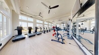 Located at 6741 Boulder Hwy in Las Vegas, NV, this well-appointed fitness center offers a comprehensive exercise environment across a single expansive floor. The space is divided into distinct workout zones, each designed to support a variety of fitness routines. Soaring ceilings and floor-to-ceiling windows along the perimeter flood the interior with natural light, providing open views of the surrounding neighborhood and creating an inviting, airy atmosphere. Multiple large ceiling fans are distributed throughout, ensuring consistent airflow and comfort during workouts. The cardio zone features a row of treadmills, stationary bikes, and elliptical machines, all positioned to take advantage of the natural light. A dedicated strength training area includes a robust selection of cable machines, multi-functional weight stations, and weight benches, transitioning into a free weights section stocked with a full dumbbell rack. Rubber flooring in the strength zones provides safety and durability, while light hardwood-style flooring in the open areas lends a clean, modern aesthetic. Wall-mounted mirrors and mounted flat-screen TVs enhance the space's functionality and ambiance. The layout flows intuitively, offering ample room for movement between zones, making this fitness center a well-equipped and comfortable facility for a full range of exercise needs.