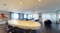 This thoughtfully designed single-floor lounge space offers a sophisticated environment suited for socializing and relaxation. At its center sits a striking curved, high-gloss white counter with bar-height seating, serving as the focal point of the open layout. The flooring throughout is rich, dark hardwood, providing a warm contrast to the cool blue-and-white color palette that defines the space.

Multiple distinct seating zones are arranged throughout the room. One area features plush navy blue sofas and armchairs accented with gold and yellow throw pillows, anchored beneath a sculptural drum pendant light. A second lounge zone offers upholstered booth-style seating in deep teal leather, accompanied by woven accent chairs and gold pendant lighting. Large-scale abstract blue artwork adorns the walls, reinforcing the coastal aesthetic.

A sleek wet bar lines one wall, featuring white cabinetry, stone countertops, glass floating shelves, stainless steel refrigeration units, and a wall-mounted television. Delicate linear pendant fixtures illuminate this area elegantly.

Floor-to-ceiling sliding glass doors flood the space with natural light and open directly onto an outdoor terrace furnished for additional seating. Sheer white curtains soften the transitions between interior and exterior, contributing to the airy, upscale ambiance throughout.