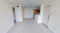 This single-floor residence offers a clean, functional layout designed for comfortable everyday living. The main living area is finished with plush, light-toned carpeting that extends throughout, creating a cohesive and welcoming atmosphere. Neutral white walls provide a bright, airy feel, enhanced by recessed lighting and a central ceiling fixture that ensures the space is well-lit throughout the day.

A defining feature of the layout is the pass-through kitchen, which opens directly into the living area via a raised breakfast bar counter. The kitchen is equipped with warm-toned wood cabinetry that adds a touch of classic character to the otherwise neutral palette. This open connection between the kitchen and living space promotes an easy flow ideal for both daily routines and casual entertaining.

The living area is flanked by multiple doorways, offering access to additional rooms and closet storage, contributing to the overall practicality of the floor plan. Ceiling vents are thoughtfully integrated throughout, indicating a central HVAC system for year-round climate comfort. The overall design is straightforward and functional, providing a versatile canvas that can be easily personalized to suit a variety of living arrangements.