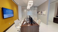 This single-floor workspace presents a sleek, contemporary conference room designed for productivity and collaboration. The centerpiece is a rich, dark walnut conference table that comfortably seats multiple participants, flanked by white ergonomic chairs with chrome frames that lend a clean, professional aesthetic throughout the space.

Multiple desktop computer workstations are integrated directly into the table, making this room well-suited for collaborative digital work or presentations. A large flat-screen monitor is mounted on the bold yellow accent wall to the left, providing a vibrant pop of color against the otherwise neutral palette of whites and grays.

A statement tiered chandelier with cascading crystal-like strands hangs above the table, serving as a dramatic focal point while providing warm, ambient lighting complemented by recessed ceiling fixtures. The back wall features translucent glass panel partitions with a subtle textured pattern, allowing diffused natural light to filter into the room while maintaining privacy.

Abstract wall art adorns the right side wall, adding a refined artistic touch. The light-toned flooring and neutral gray walls create a bright, open atmosphere, contributing to an overall ambiance that is both functional and visually sophisticated.
